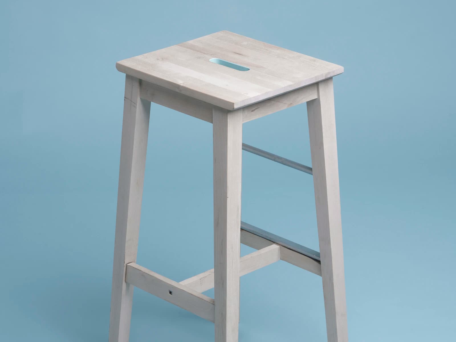 Editorial photograph of a pale solid-wood stool against a blue background